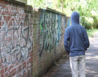 Photo showing a teenage boy / youth wearing a blue hoodie and standing beside a brick wall in an overgrown alleyway, in a run-down part of the city.